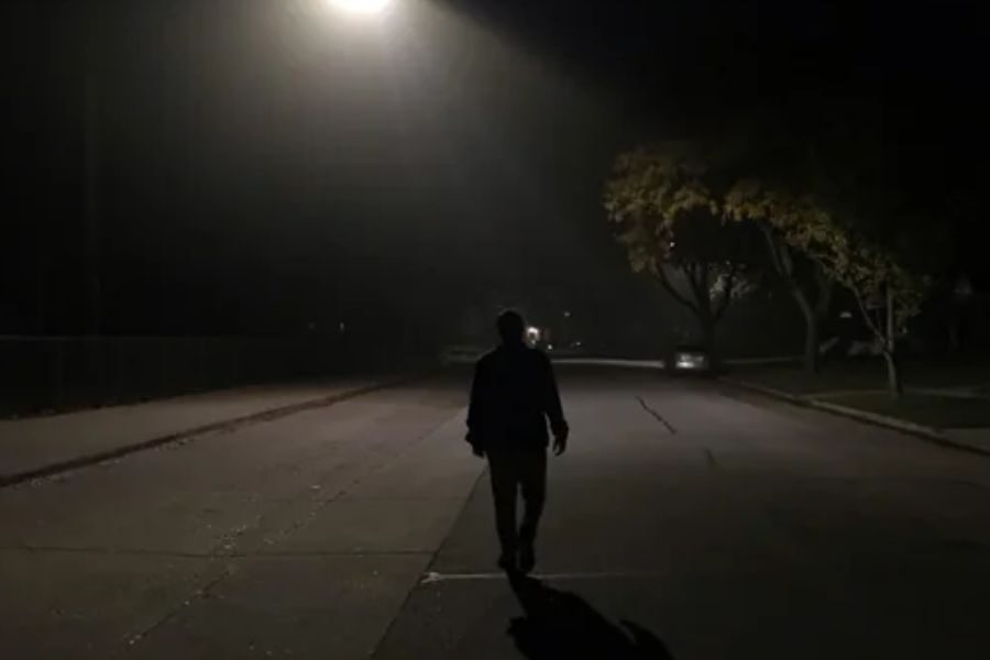 Silhouette of a person walking down a dimly lit suburban street at night under a single streetlamp.