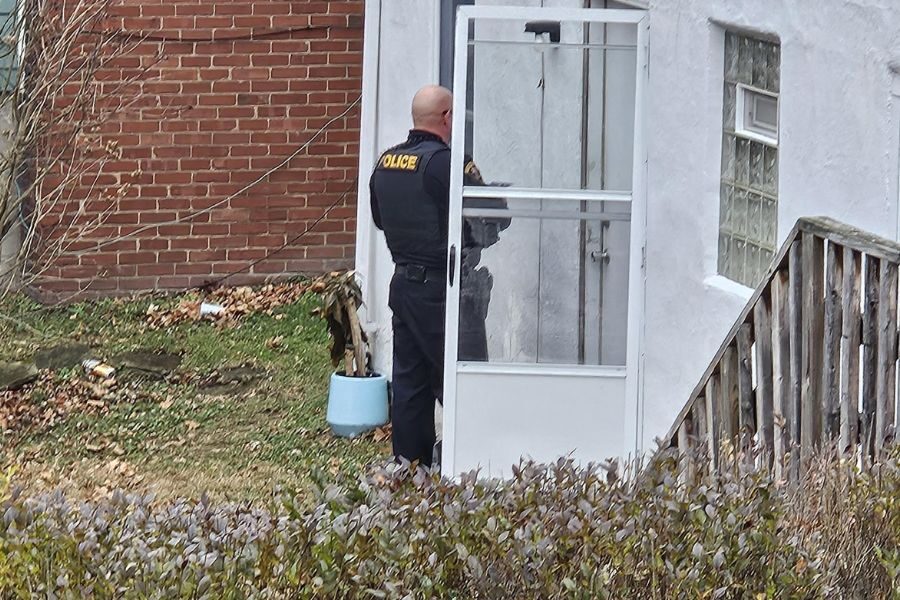 A police officer in uniform stands at the entrance of a white house, speaking to someone through the screen door.
