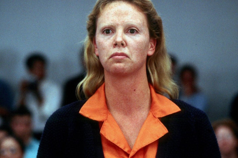 A woman with pale, makeup-free skin and a stern expression wears an orange prison uniform and dark sweater, standing in a courtroom with a blurred crowd behind her.
