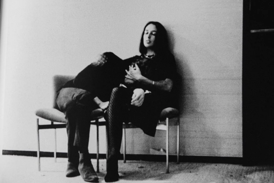 Black and white photo of Joan Baez sitting in a chair with Bob Dylan lying across her lap, his head resting on her chest as she gently holds him.
