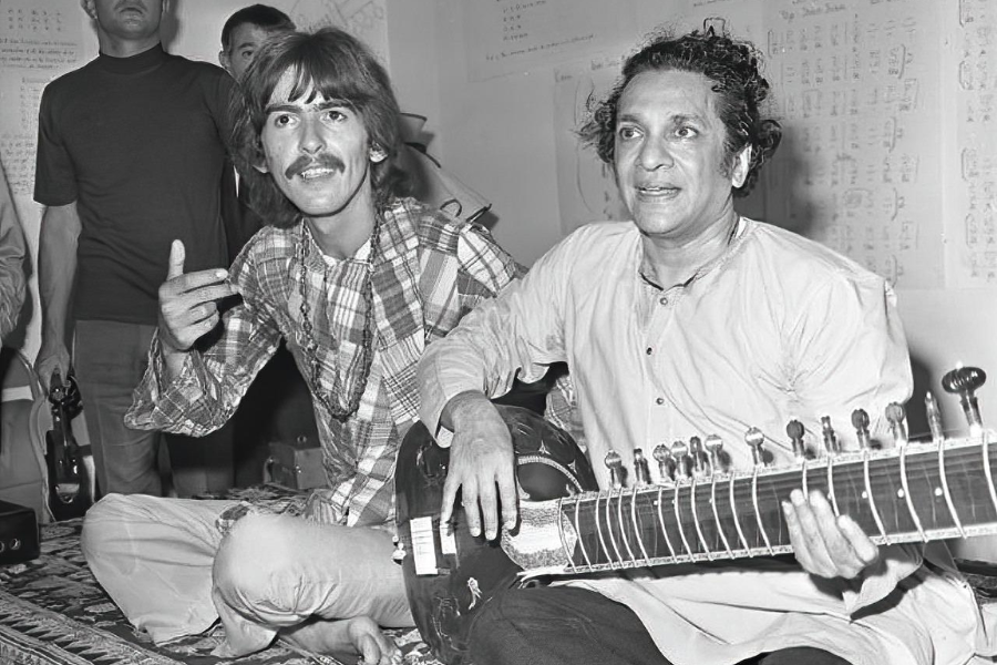 George Harrison sits cross-legged next to sitar maestro Ravi Shankar, smiling and gesturing with his hand during a lesson.