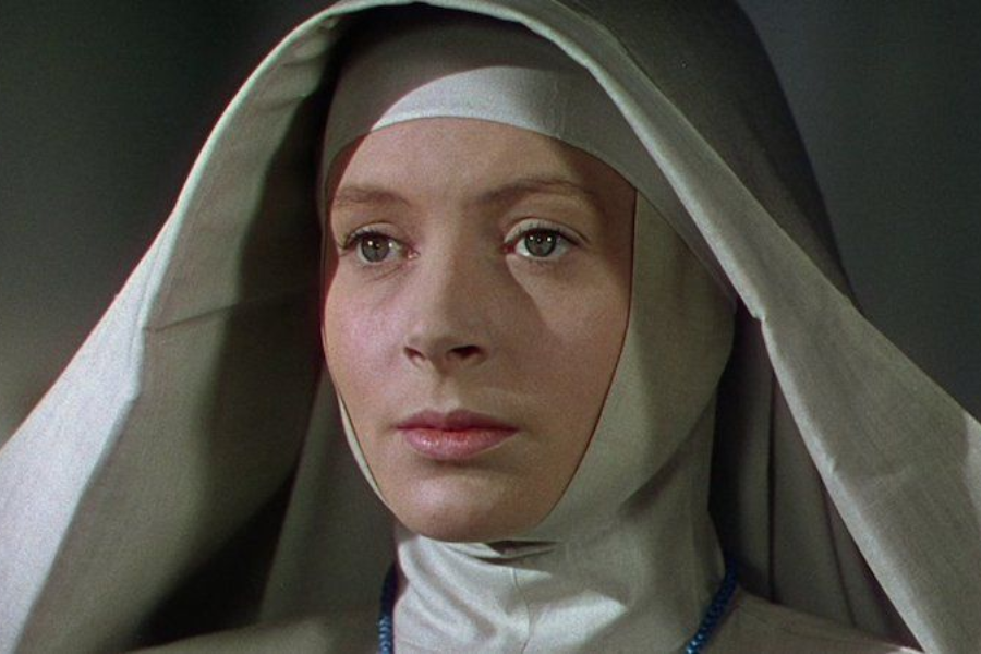 A close-up of a woman dressed as a nun in a light grey habit, with a calm, solemn expression and a soft focus background.