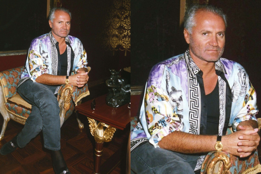 Seated man wearing a colorful silk shirt with Greek key trim and bold prints, paired with black jeans and gold accessories. Close-up shows his composed expression and luxurious surroundings.