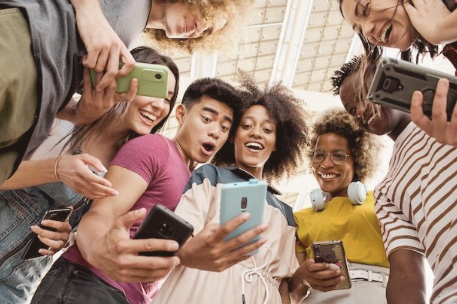 A diverse group of young people huddle together smiling and laughing as they look at smartphones, clearly enjoying a shared moment.