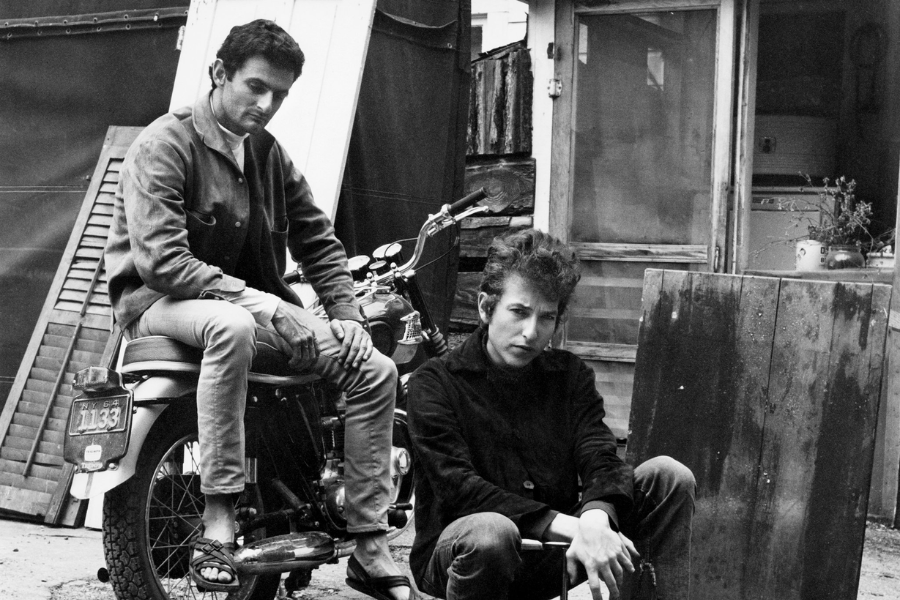 Black and white photo of Bob Dylan sitting on the ground beside a man perched on a motorcycle in a rustic outdoor setting.
