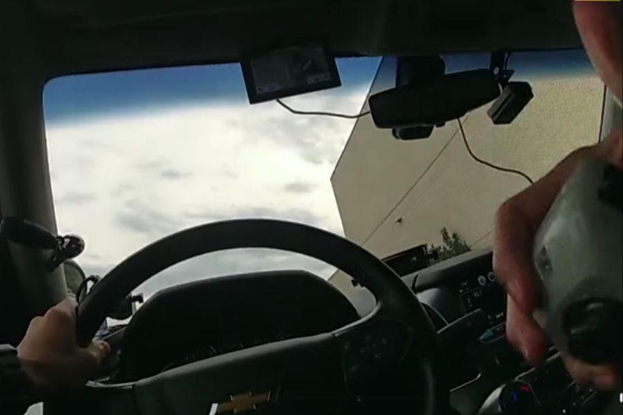 View from inside a police vehicle showing a hand on the steering wheel and another holding a device, with a building visible through the windshield.