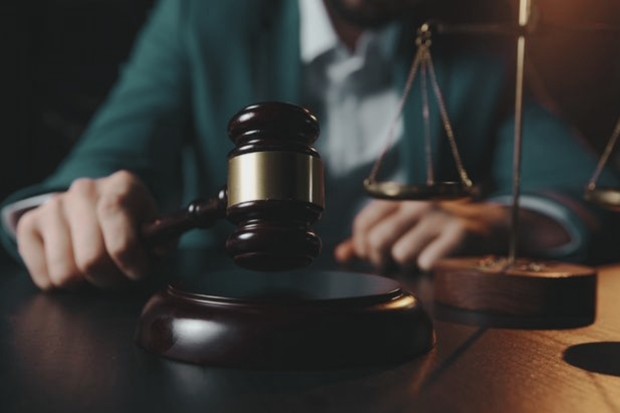 A judge's gavel is raised mid-motion beside a scale of justice, with a person in a dark green jacket blurred in the background.