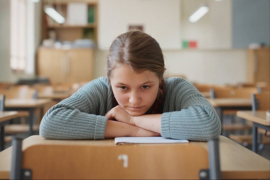A young girl sits alone in an empty classroom with her arms crossed on a desk, staring down at a notebook with a tired expression.