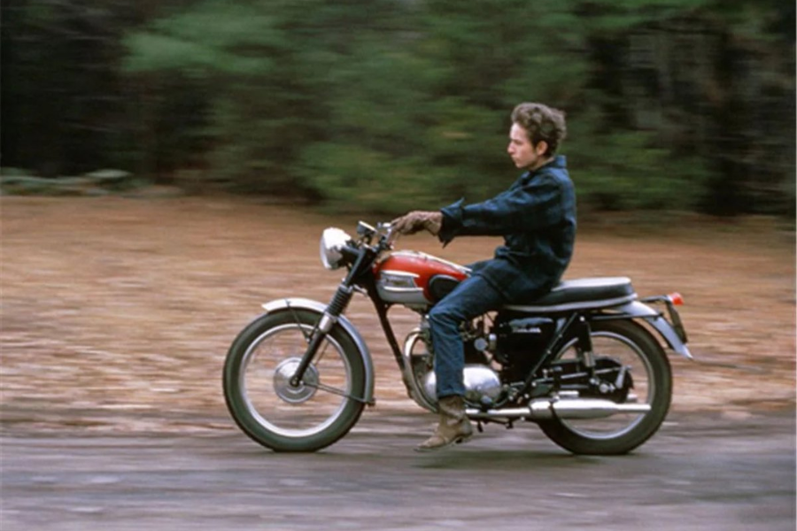 A young Bob Dylan rides a red and silver Triumph motorcycle on a road surrounded by trees and autumn leaves.