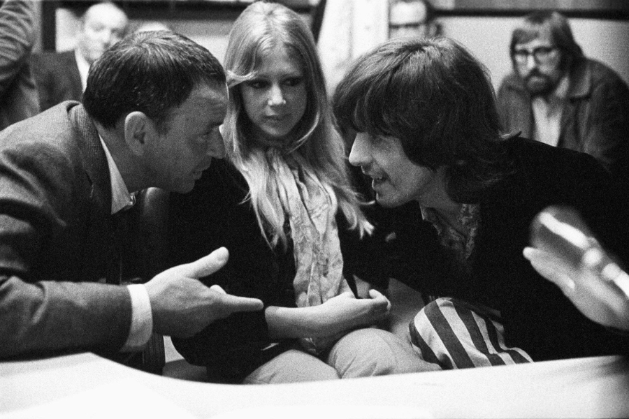 George Harrison and Pattie Boyd sit in an intense conversation with a man gesturing while speaking to them during a public appearance.