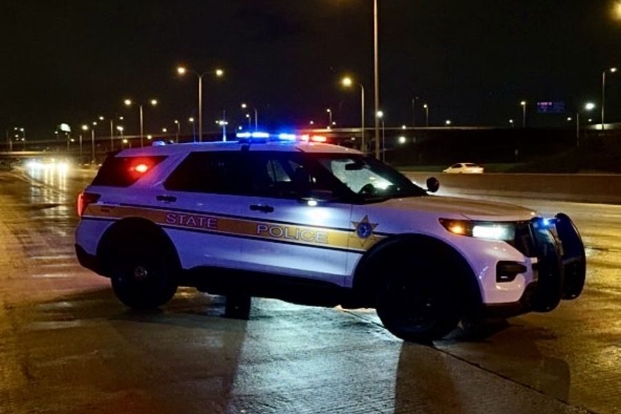 A state police SUV with flashing lights parked across a wet highway at night, partially blocking traffic.