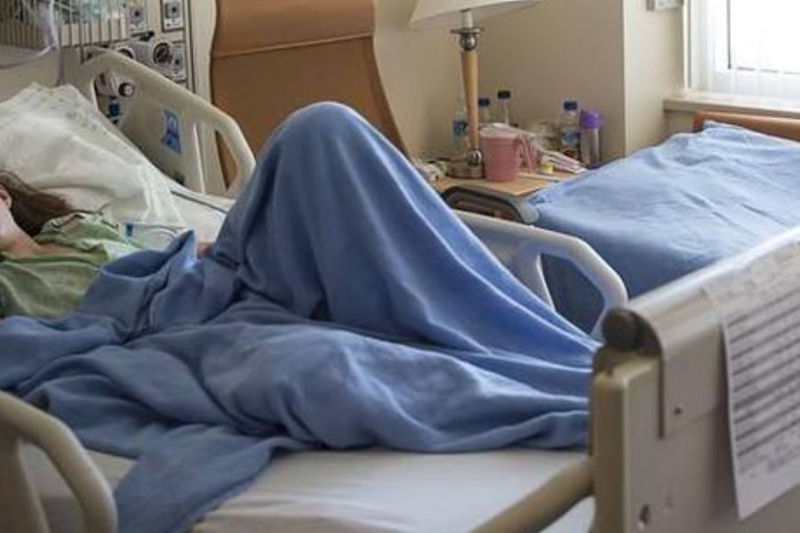 Hospital room with a patient lying under a blue blanket in a bed, and another bed nearby with items on a table between them.