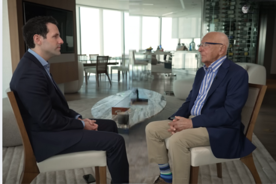 Two men in suits sit across from each other in a modern, bright room during a formal interview, with a coffee table and open space between them.