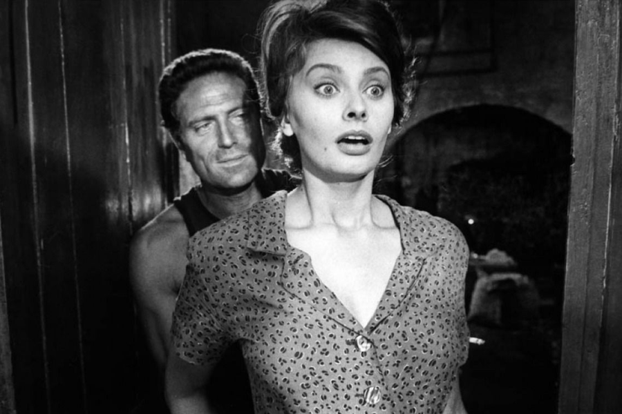 In a black-and-white image, a man stands closely behind a wide-eyed woman who appears startled, wearing a patterned dress in a dimly lit setting.