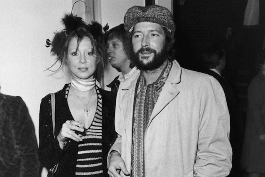 Pattie Boyd wears a striped dress and scarf while standing beside Eric Clapton, who is dressed in a cap and coat at a 1970s event.