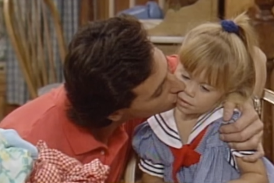 A man kisses a little girl on the cheek in a tender moment from a scene on "Full House." The girl wears a blue sailor dress with a red ribbon and looks slightly indifferent.