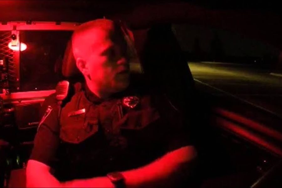 A police officer illuminated by red light sits in the driver’s seat of a patrol car at night, looking to the side.