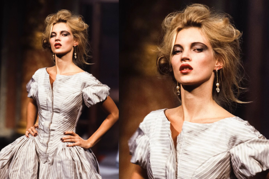 Model with voluminous tousled hair and pearl earrings poses in a corset-inspired off-white gown with subtle stripes, hands on hips. Close-up highlights her striking makeup and confident expression.