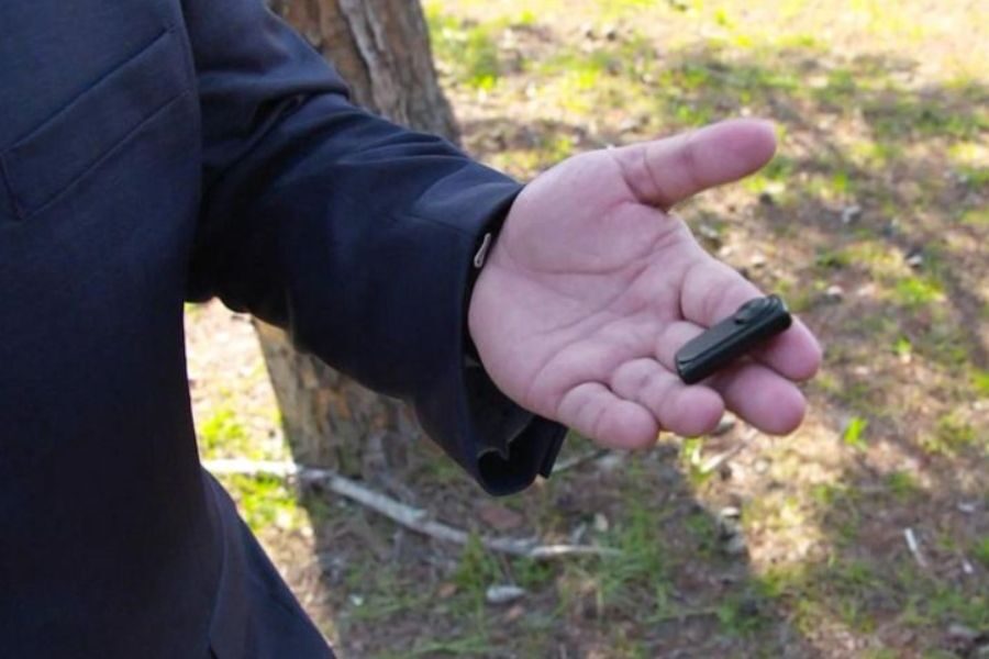 A close-up of a man's hand holding a small black voice recorder, with a blurred wooded area in the background.