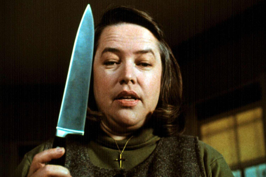 A woman stares at a large kitchen knife she's holding upright, with a blank expression and wearing a dark sweater and gold cross necklace.