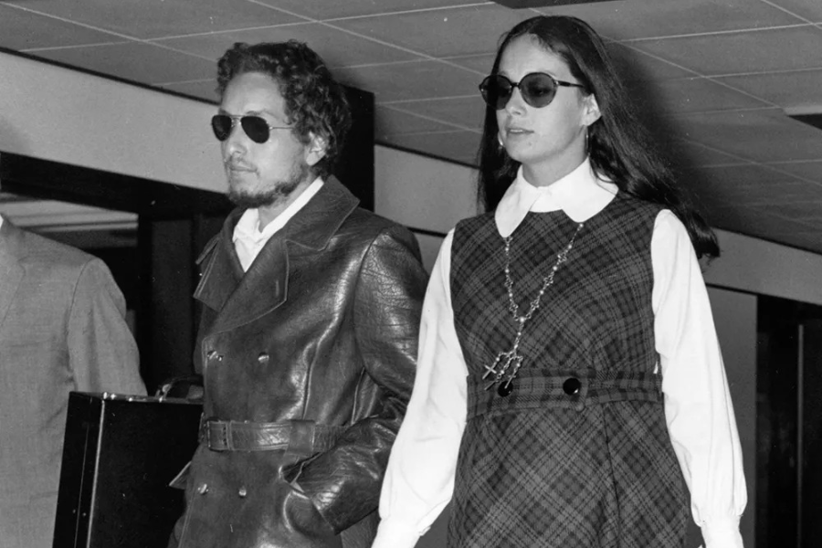 Black and white photo of Bob Dylan and Sara Dylan walking through an airport wearing sunglasses, with Dylan in a leather trench coat.