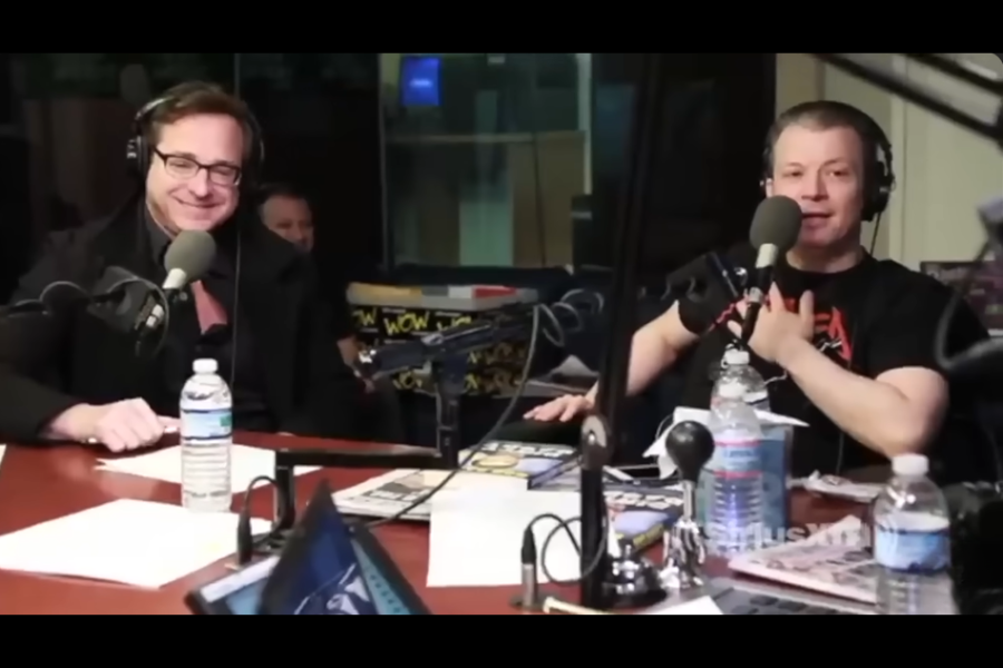 Two men sit at a podcast-style table with microphones, headphones, and water bottles during a radio broadcast. One man smiles while the other speaks into the mic.