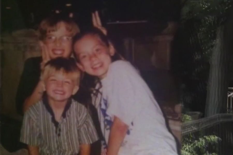 Three smiling children pose for a photo, with one older girl flashing bunny ears behind a boy's head in a playful gesture.