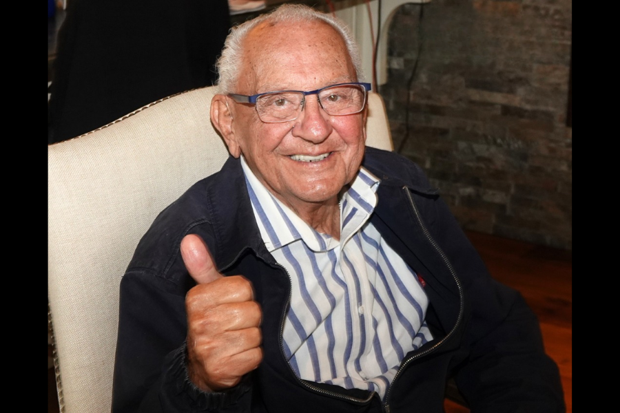An older man, smiling broadly, gives a thumbs-up gesture while sitting indoors, wearing a striped shirt and glasses.