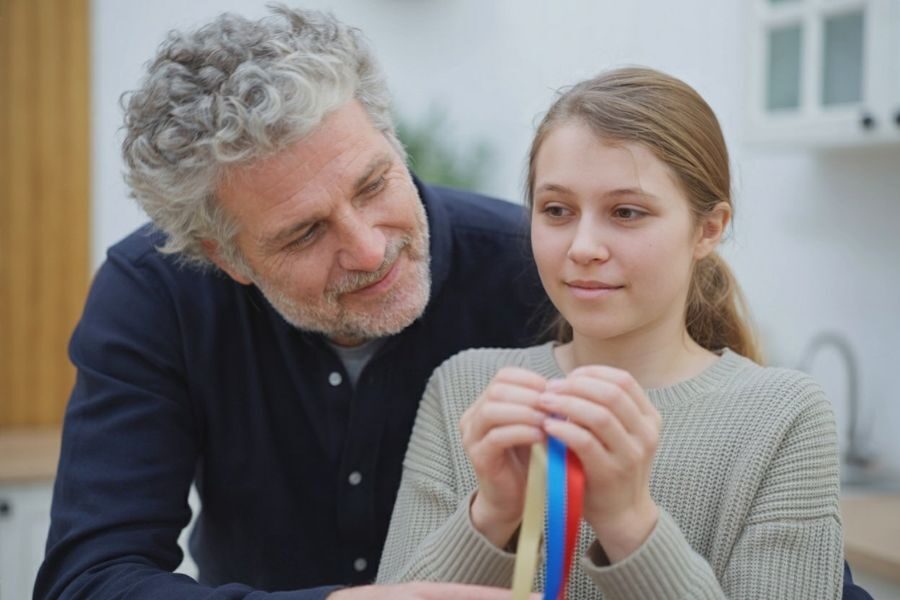 A smiling man looks warmly at a girl holding colorful award ribbons, sharing a proud and affectionate moment.