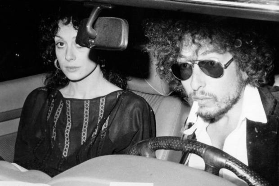 Black and white image of Bob Dylan driving a car at night with a woman in the passenger seat looking directly at the camera.