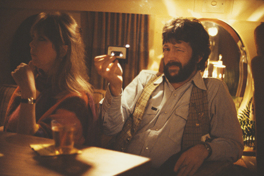 Pattie Boyd and Eric Clapton sit aboard a private jet, with Clapton holding up a cassette tape and squinting at the camera.
