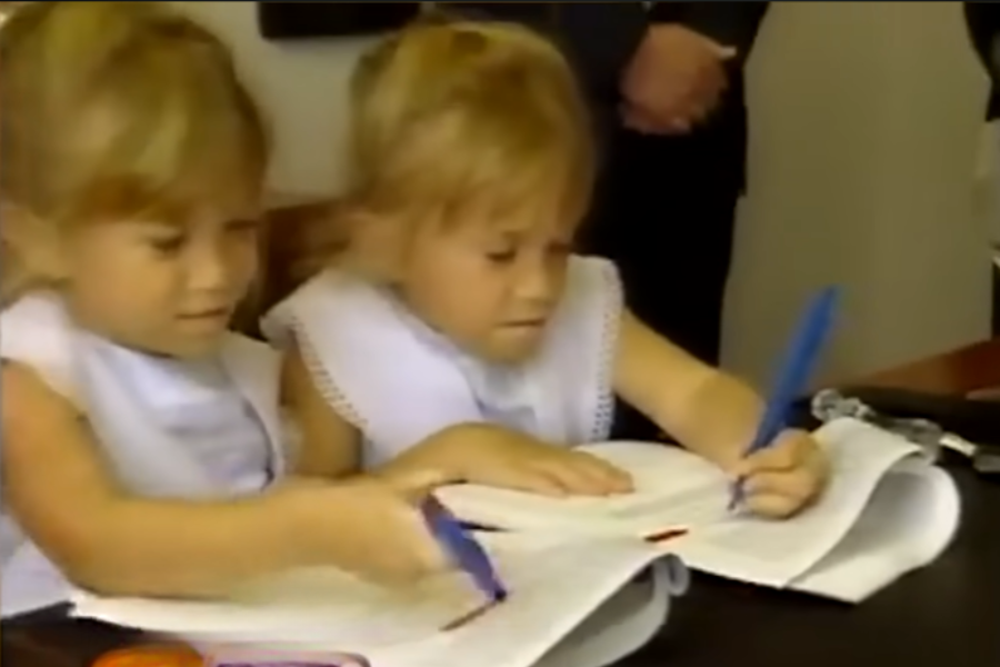 Twin girls, both wearing white sleeveless dresses, focus intently while signing documents with oversized pens. The scene appears to be a formal or promotional signing.