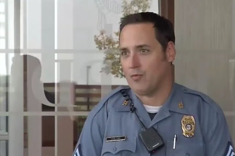 olice officer in a blue uniform stands outside a building, mid-sentence during an interview or conversation.