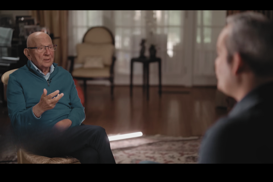 An older man speaks while gesturing with his hand during a one-on-one interview in a warmly lit living room, facing an off-camera host.