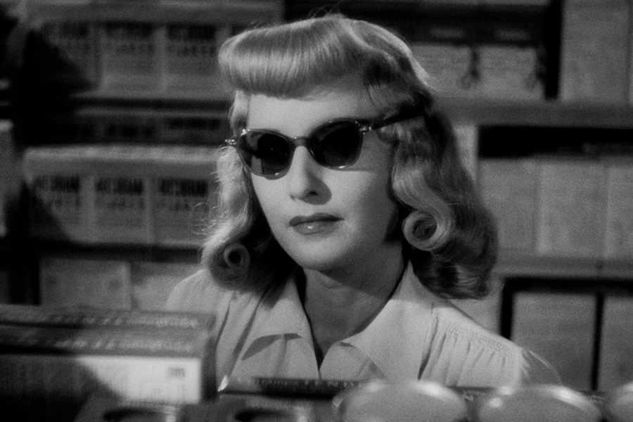 In a black-and-white film still, a woman with curled blonde hair and dark sunglasses stands near shelves, exuding mystery and confidence.