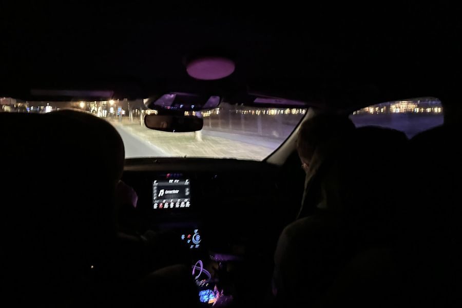 A nighttime view from inside a moving vehicle, showing two passengers and city lights through the windshield.