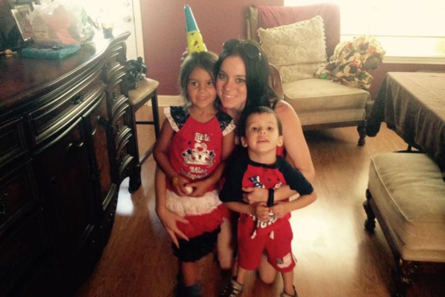 Woman kneeling and hugging two young children inside a home, with the girl wearing a birthday hat and the boy smiling at the camera.