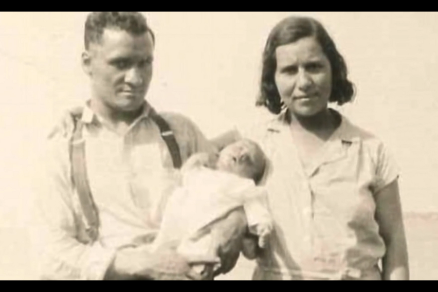 A black-and-white photo shows a man and woman holding a baby, dressed in early 20th-century attire, standing outside in natural light.