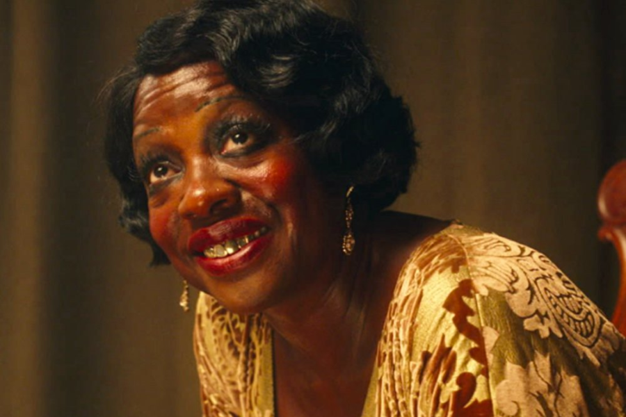A woman in gold patterned clothing with bold red lipstick and exaggerated makeup smiles up from her seat, wearing statement earrings.
