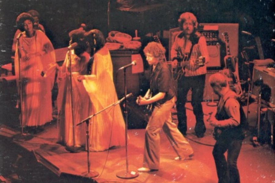 Bob Dylan performing on stage with a full band and backup singers, all bathed in reddish stage lighting during a live concert.
