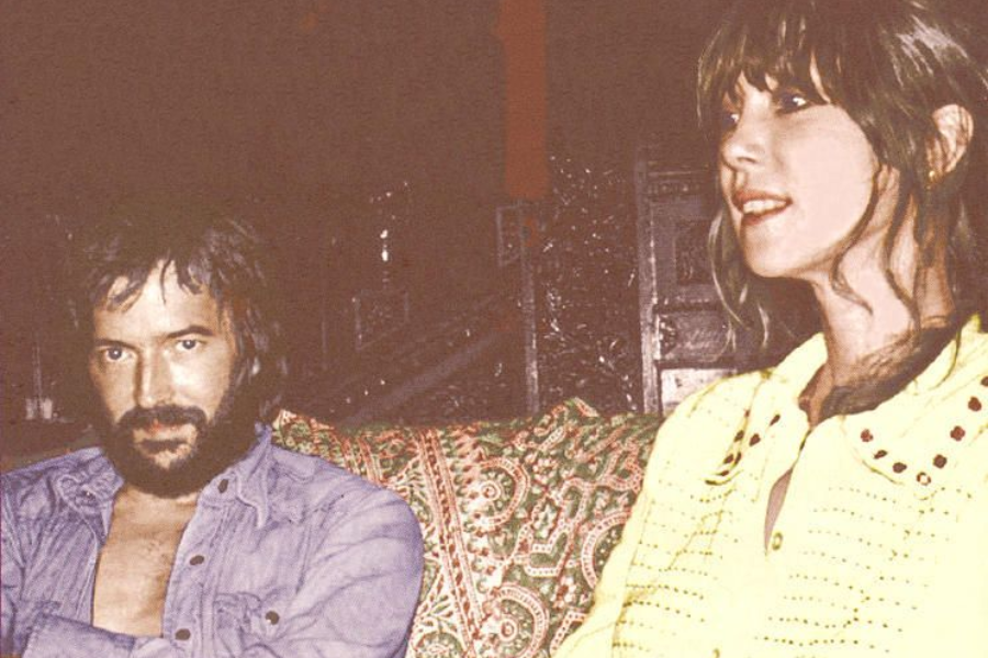 Eric Clapton and Pattie Boyd sit on a couch with intricate fabric, Clapton gazing toward the camera while Boyd looks off to the side.