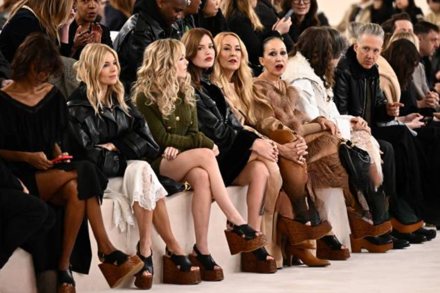 A group of fashionably dressed people sit in the front row at a fashion show, many of them wearing bold platform shoes, chatting and watching the runway.