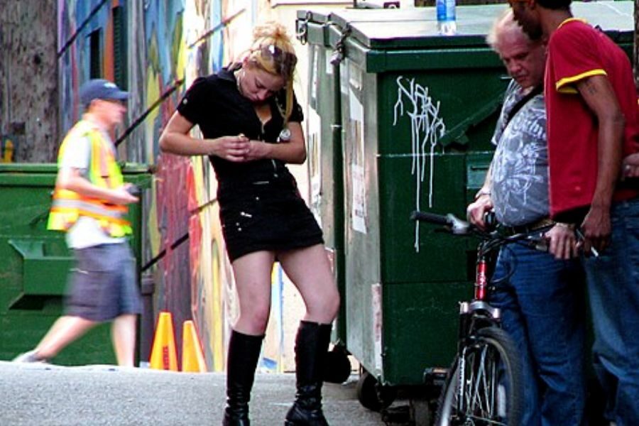 A woman in a short black outfit stands beside two men and a bike near a graffiti-covered alley with dumpsters.
