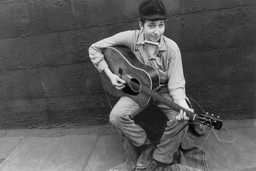 A black and white image of a young man sitting casually with a guitar and harmonica, leaning forward and smiling slightly at the camera.