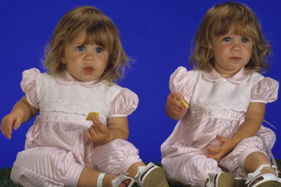 Toddler twins sit against a bright blue backdrop, each holding a snack, dressed in light pink and white puff-sleeve outfits with white sandals.