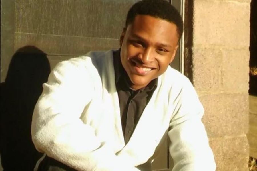 A smiling young man in a white cardigan and black shirt, sitting outside in warm sunlight.