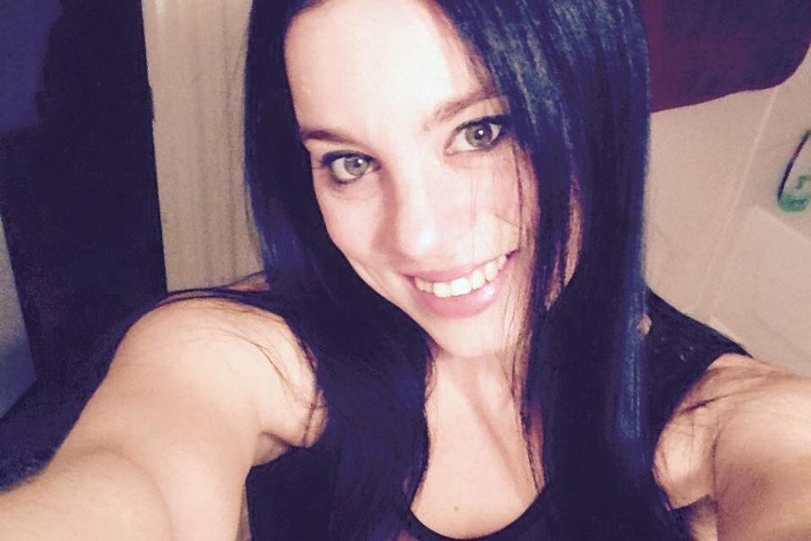 Close-up selfie of a woman with long black hair and green eyes, smiling in a well-lit bathroom.