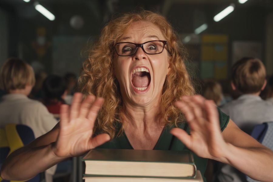 A red-haired teacher with glasses screams in shock or frustration, hands raised in front of a stack of books in a classroom.