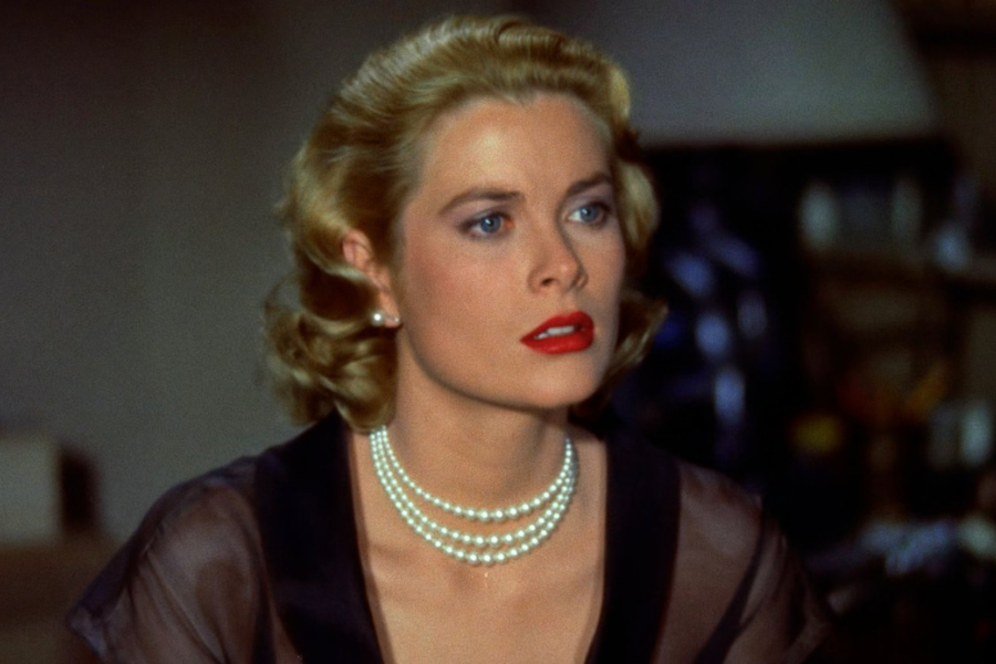 A poised woman with red lipstick and blonde curled hair looks concerned, wearing a sheer black top and layered pearl necklace in a softly lit room.