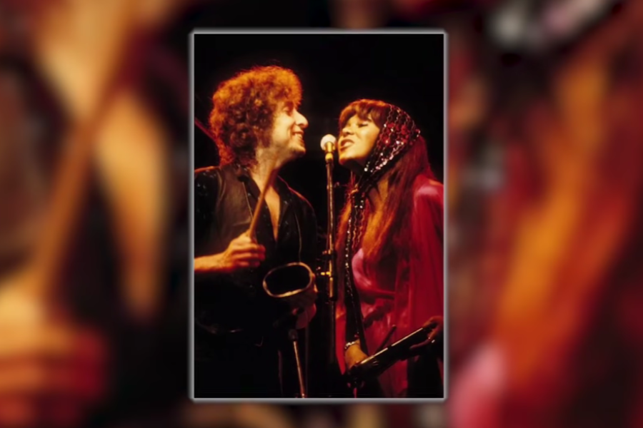 Bob Dylan and a female vocalist smiling at each other as they share a microphone on stage, captured mid-performance with vibrant lighting.